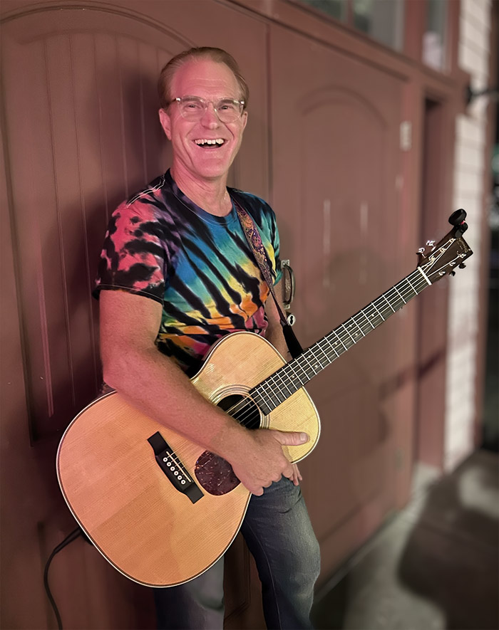 Tom, wearing a colorful tie-die shirt, standing with guitar, smiling.